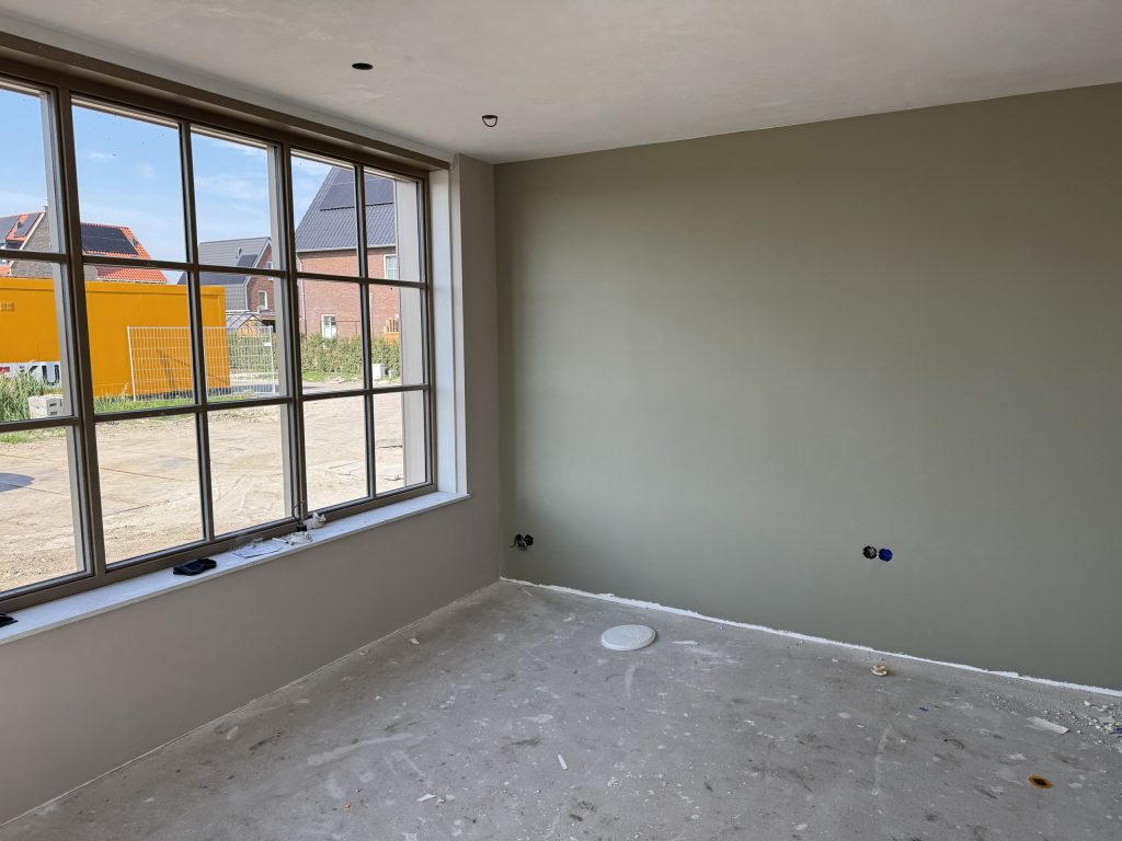 Nieuwbouwkamer met een grijsgroen gesausde wand en een groot raam met uitzicht op een bouwterrein