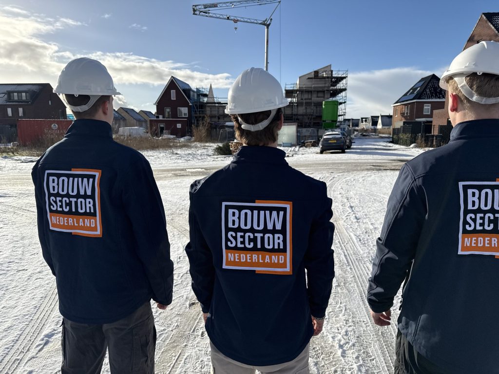Drie vakmensen van Bouwsector Nederland in donkere werkjas en witte helm kijken naar nieuwe huizen